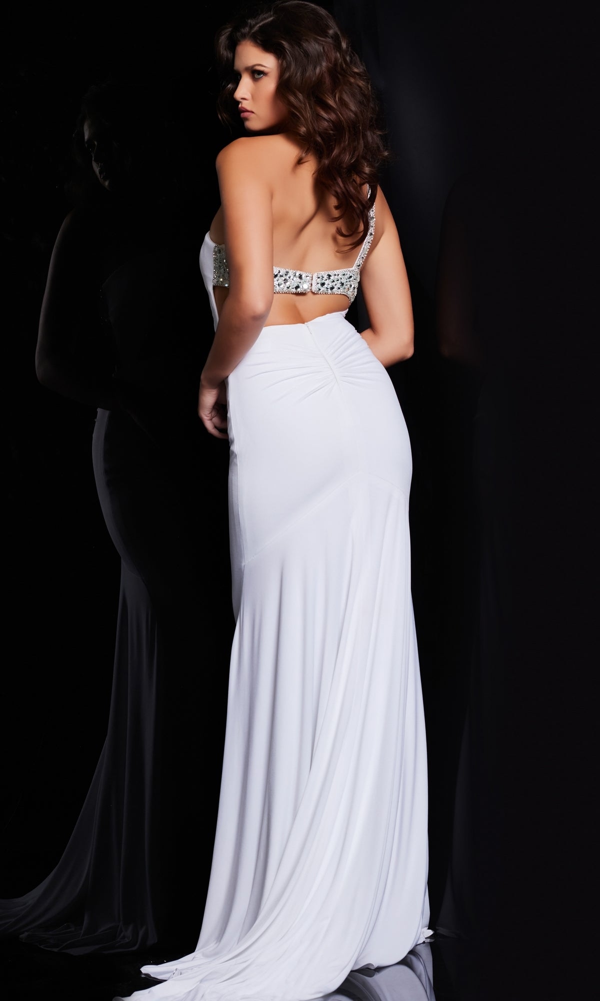 A woman with wavy brown hair models the Jovani Cut-Out Beaded-Bodice Long Prom Dress 23130, featuring jeweled details and an open back, against a dark background.